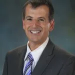 A man named Geoff Gutgold is smiling for the camera, wearing a gray suit with a white shirt and a striped tie. The background features a soft gradient of blue and green hues.