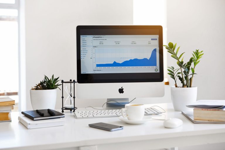 A desktop computer displaying a blue graph on its screen is positioned on a white desk. Next to the computer, there is a white coffee cup, a smartphone, a stack of books, and two potted plants. This workspace suggests a focus on marketing analytics.