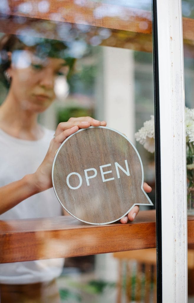 A person placing a round wooden "OPEN" sign on a glass door of a shop, indicating the business is open. The setting includes wooden decor and bright natural light, suggesting a welcoming environment for small business advertising.