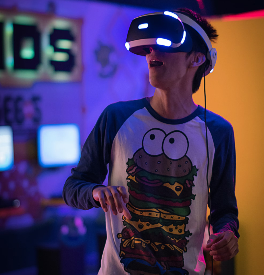 A young man wearing a virtual reality headset and a shirt featuring a cartoon hamburger stands in a dimly lit arcade. The background shows arcade machines and bright colors, creating a playful gaming environment.