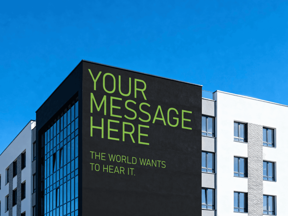 A modern building features a large black wall displaying bright green text that reads "YOUR MESSAGE HERE" and below it, "THE WORLD WANTS TO HEAR IT". The clear blue sky serves as the backdrop, highlighting the regional advertising potential.