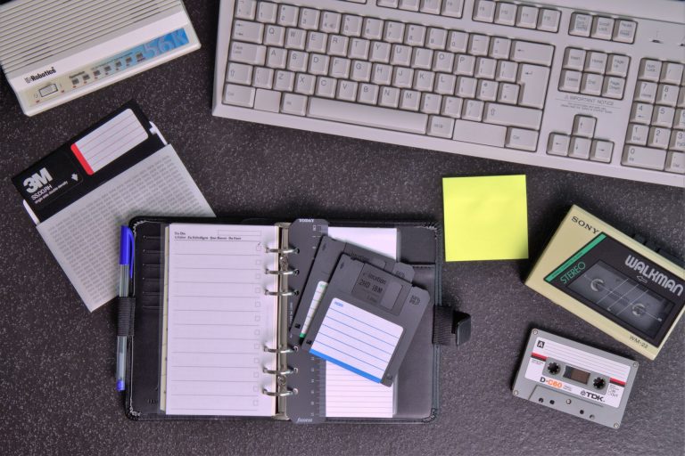 A black organizer with opened pages sits on a dark desk, accompanied by blue and black pens, paper notes, floppy disks, a yellow sticky note, and a Sony Walkman cassette player. A gray computer keyboard is partially visible in the background, reflecting the setting of an advertising agency.